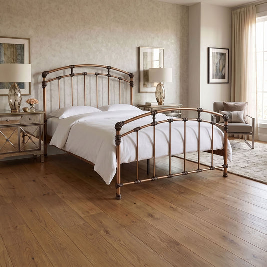 Aristella Iron Bed by Wesley Allen in Aged Brass finish in Queen size. A metal bed frame with an arched headboard and footboard in a warm Aged Brass, two-tone finish, styled in a bright, modern bedroom with light wood floors, white bedding, and matching bedside tables.