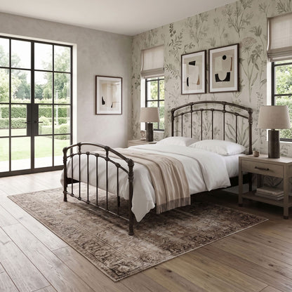 Test Iron Bed by Wesley Allen in Aged Bronze finish in Queen size. A styled bedroom interior featuring a dark metal bed frame with a curved headboard and footboard, dressed in white bedding and a beige throw. The bed is placed on a distressed brown area rug over light wood flooring, flanked by matching bedside tables and lamps.
