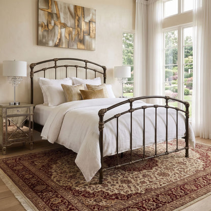 Test Iron Bed by Wesley Allen in Aged Gold finish in Queen size. A dark metal bed frame with an arched headboard and footboard, styled with white bedding and decorative pillows in a bright bedroom setting with a patterned rug and large windows.