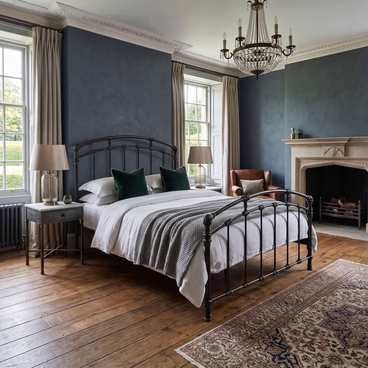 Test Iron Bed by Wesley Allen in Aged Iron finish in Queen size. A styled bedroom featuring an antique black metal bed frame with white bedding, green accent pillows, a gray textured throw, a dark blue wall, wooden floors, a fireplace, and a chandelier.