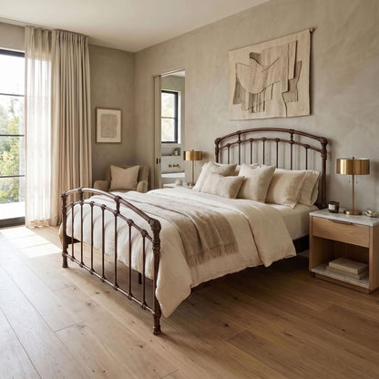 Test Iron Bed by Wesley Allen in Copper Bisque finish in Queen size platform iron frame with wood slats. A rustic dark metal bed frame with a curved headboard and footboard, dressed in cream bedding, set in a modern, neutral-toned bedroom with wood floors and a matching nightstand.