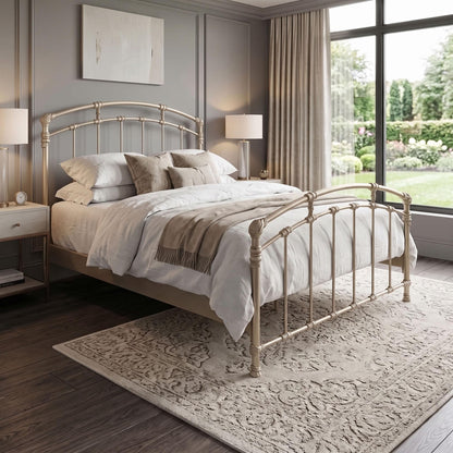 Test Iron Bed by Wesley Allen in Queen size. A classic metal bed frame in a warm Hand Brushed Champagne finish, styled in a modern bedroom setting with white and beige linens, nightstands, and a large patterned area rug.