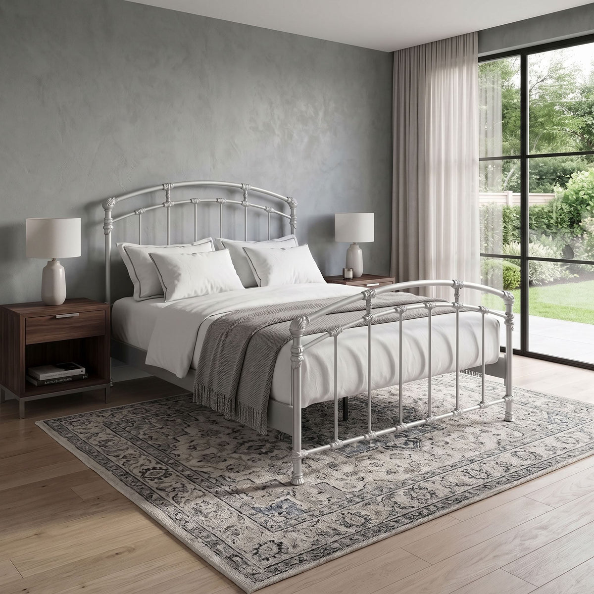 Test Iron Bed by Wesley Allen in Opaque Light Silver finish in Queen size platform frame with wood slats. A silver metal bed frame with a curved headboard and footboard, styled with white bedding and a gray throw blanket, set on a patterned area rug in a modern bedroom.
