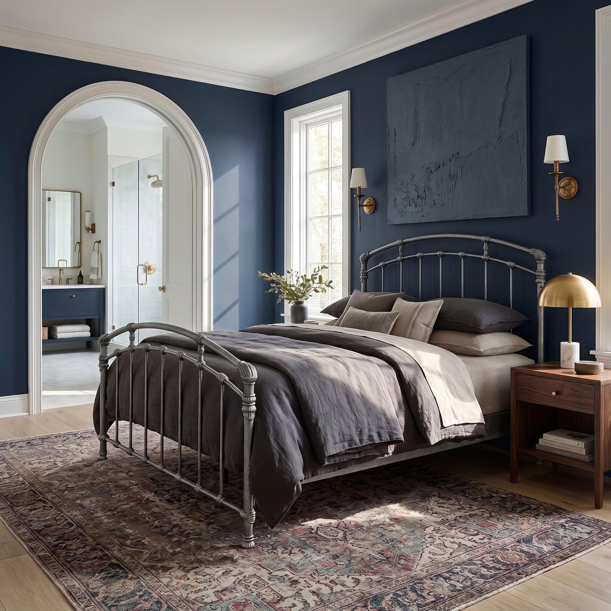 Test Iron Bed by Wesley Allen in Silver Bisque finish, Queen size, and a platform frame with wood slats. A styled bedroom interior featuring a metal bed frame with gray bedding, set on a patterned area rug against a dark blue wall. The room includes a wooden nightstand with a gold lamp, wall sconces, a large abstract painting, and an arched doorway leading to a bathroom.