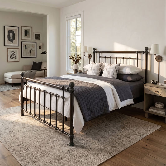 Revered Iron Bed by Matriae in the Aged Bronze finish queen size with a wood slats platform bed frame. A dark metal bed frame with turned posts and a curved headboard and footboard, styled in a bright, modern bedroom with gray and white bedding, a patterned rug, and wooden floors.