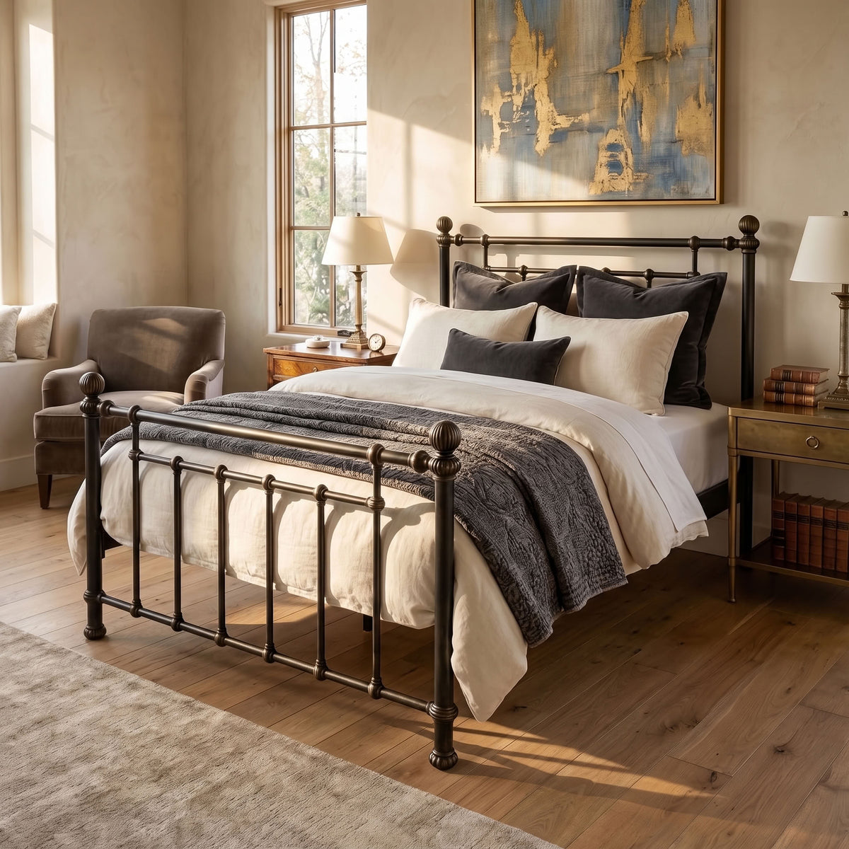 Revered Iron Bed by Matriae in the Aged Gold finish queen size with a wood slats platform bed frame. A classic metal bed frame with turned posts and a slatted guard rail, styled in a warm, sunlit bedroom with beige walls, wood floors, and neutral bedding.
