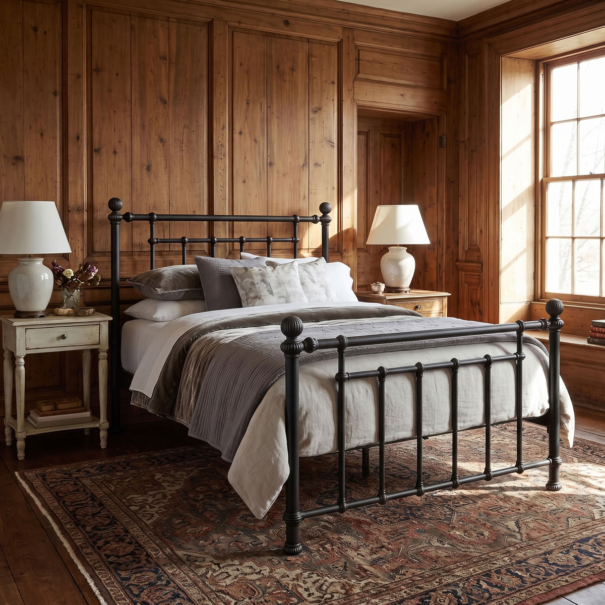 Revered Iron Bed by Matriae in the Aged Iron finish, also called antique black, queen size with a wood slats platform bed frame. A rustic bedroom scene featuring a black metal bed frame with a classic turned-post design, dressed in neutral gray and beige bedding. The bed is set in a warm wood-paneled room with matching wood nightstands, white table lamps, and a traditional patterned rug.