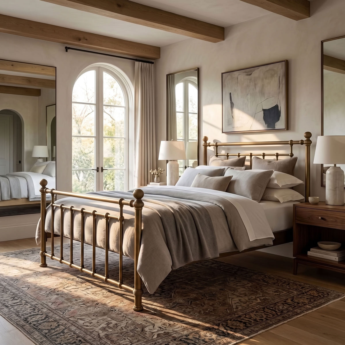 Revered Iron Bed by Matriae in the Brass Bisque finish queen size with a wood slats platform bed frame.A styled bedroom interior featuring a brass metal bed frame with a classic spindle headboard and footboard, dressed in neutral beige and gray bedding. The room has warm wood ceiling beams, arched windows with sheer curtains, a patterned area rug, and matching wooden nightstands and mirrors.